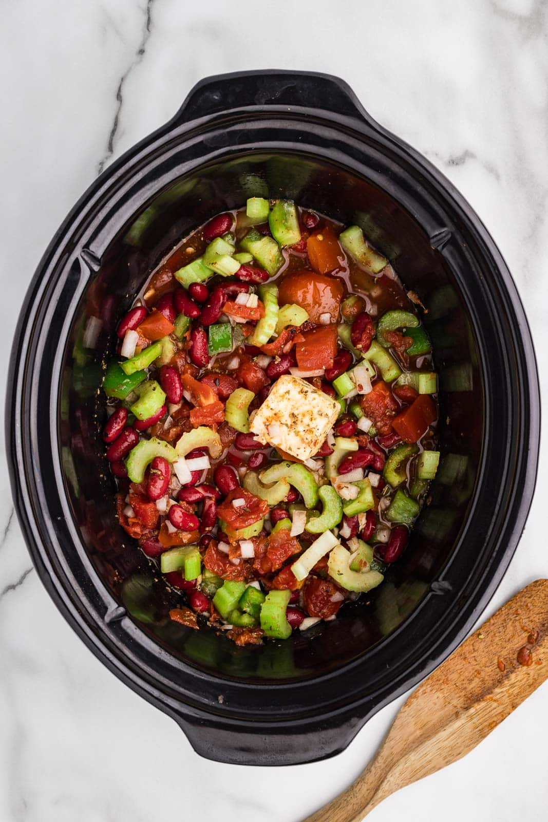 overhead view of all ingredients (except rice) stirred, in black slow cooker with wooden spoon on side