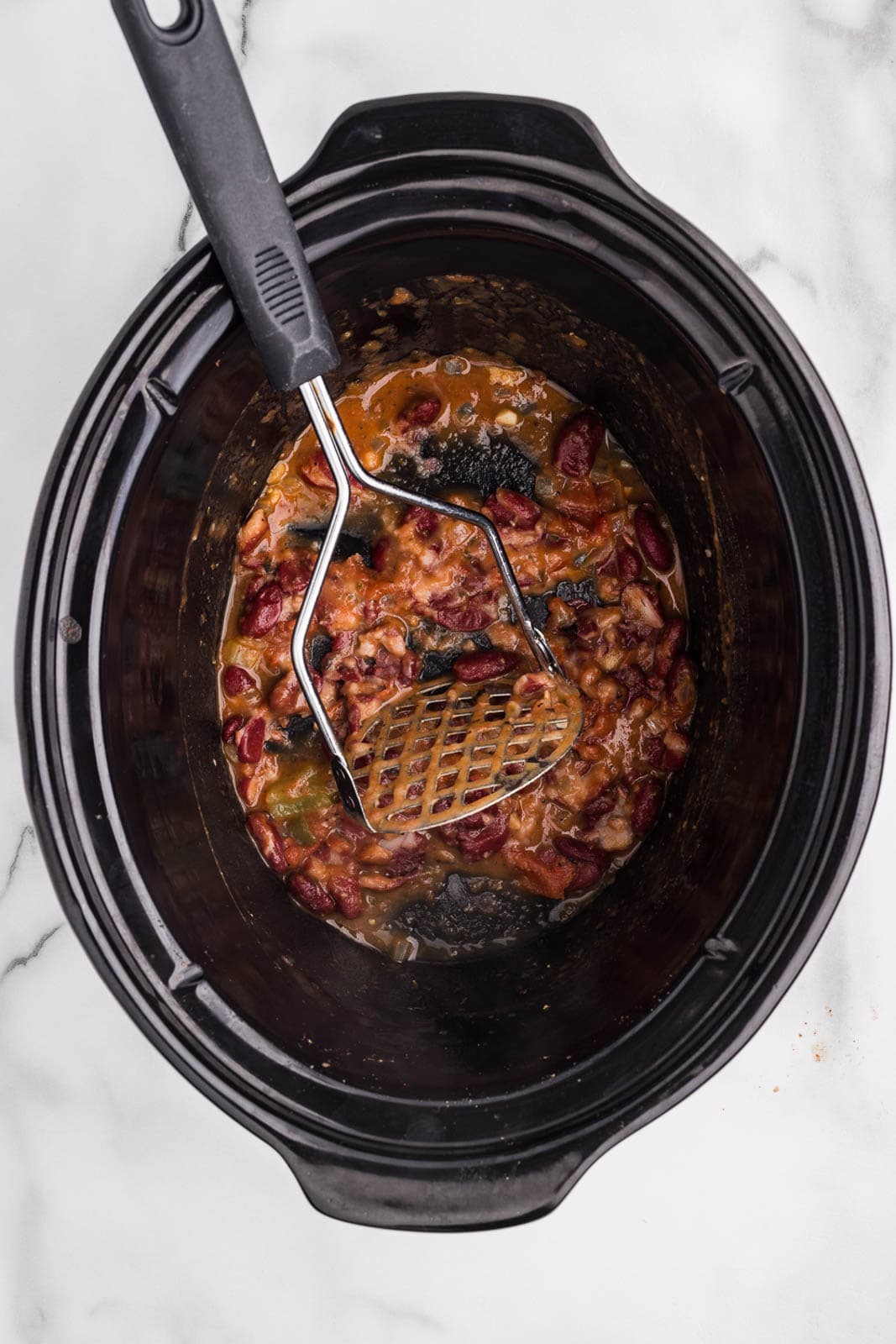 overhead view of cooked ingredients in black slow cooker with potato masher