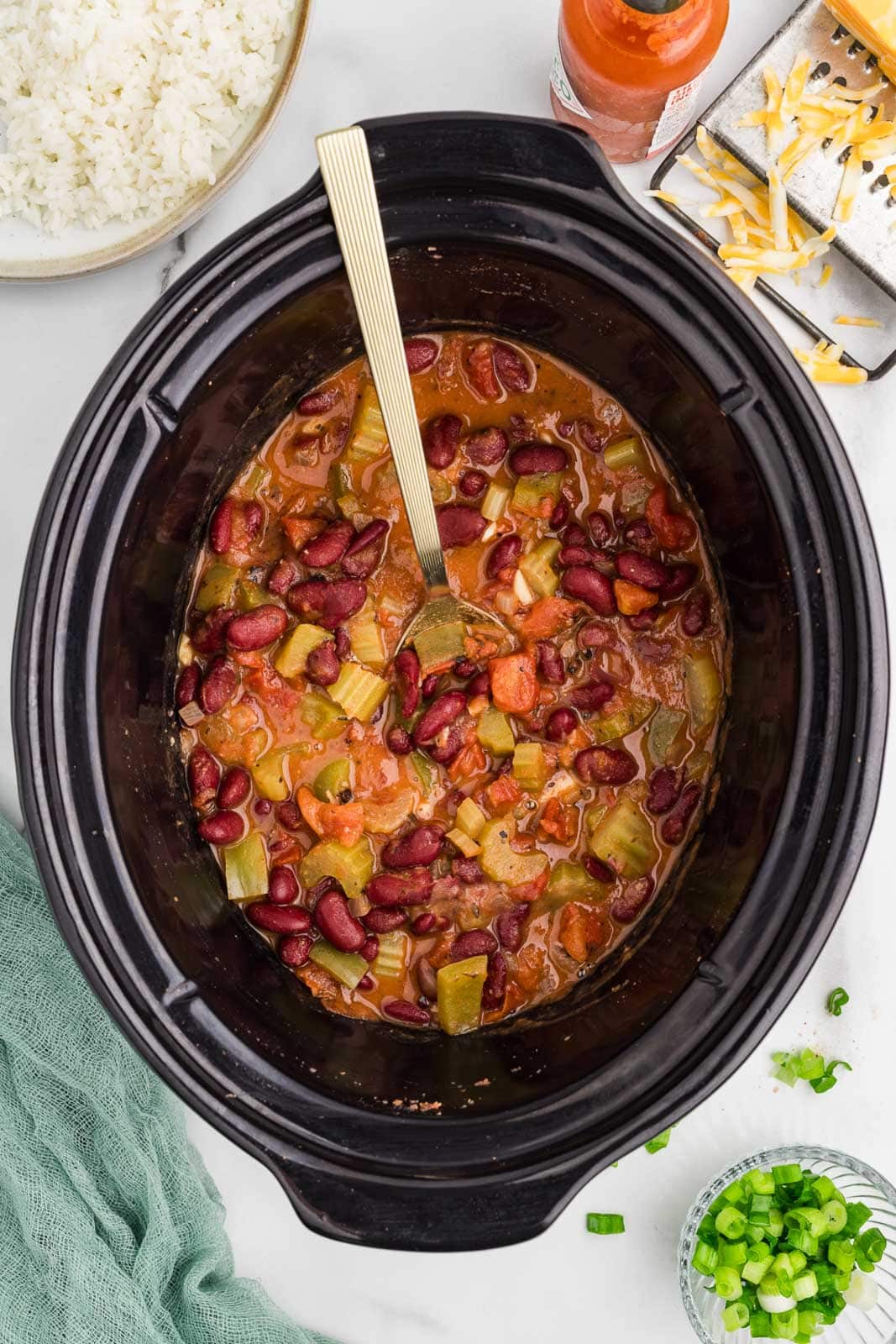 cooked red beans mixture in black slow cooker with gold spoon, with cheese shredder and cheese, hot sauce, green onions and white rice on side