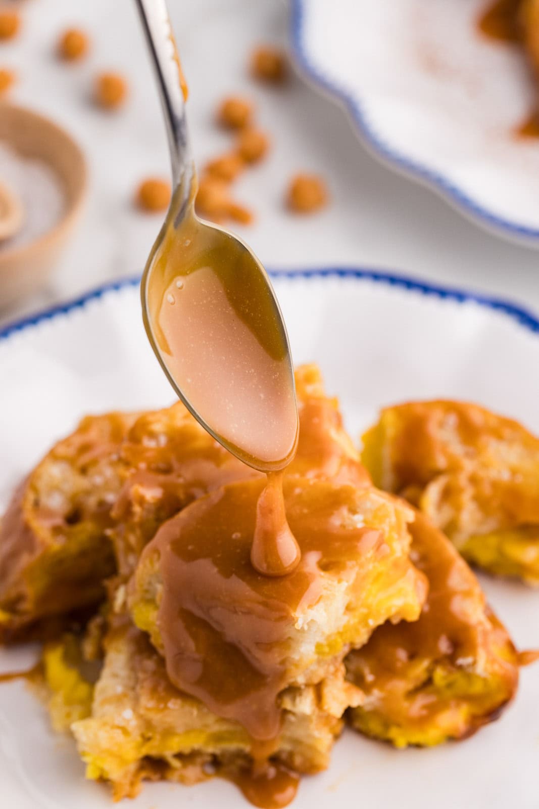 closeup of spoon with caramel mixture dropping onto french toast serving in a white plate with blue edging, another plate and salt container in background