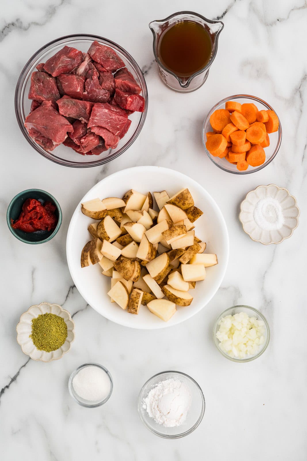 ingredients for beef stew including white bowl of cut up potatoes, white glass bowl of beef tips, glass container of beef broth, glass bowls of cut up carrots, white onion, tapioca pearls, sugar, herb seasoning, salt and colored bowl of tomato paste