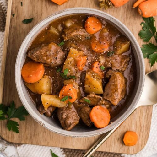 bowl of beef stew on wooden cutting board with silver spoon, raw carrots, raw potato