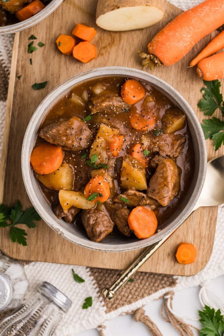 bowl of beef stew on wooden cutting board with silver spoon, raw carrots, raw potato