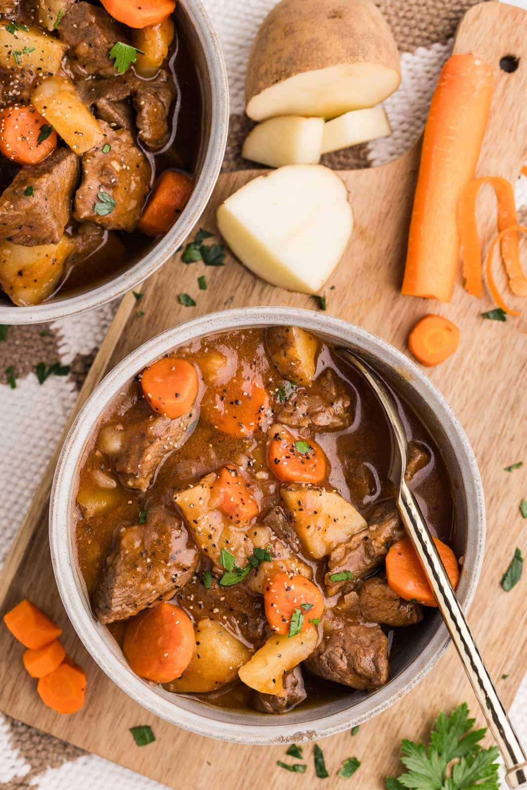 bowl of beef stew with silver spoon on a wooden cutting board with raw carrots and potatoes with another bowl in the background