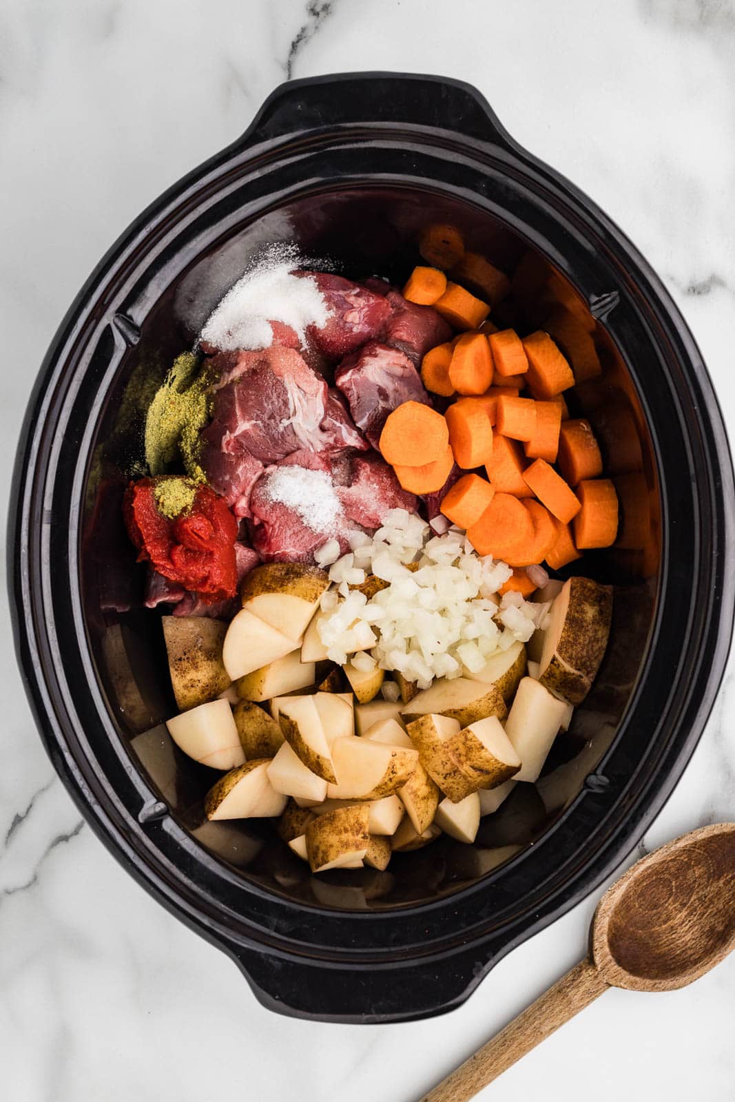 black slow cooker containing all ingredients with wooden spoon on side