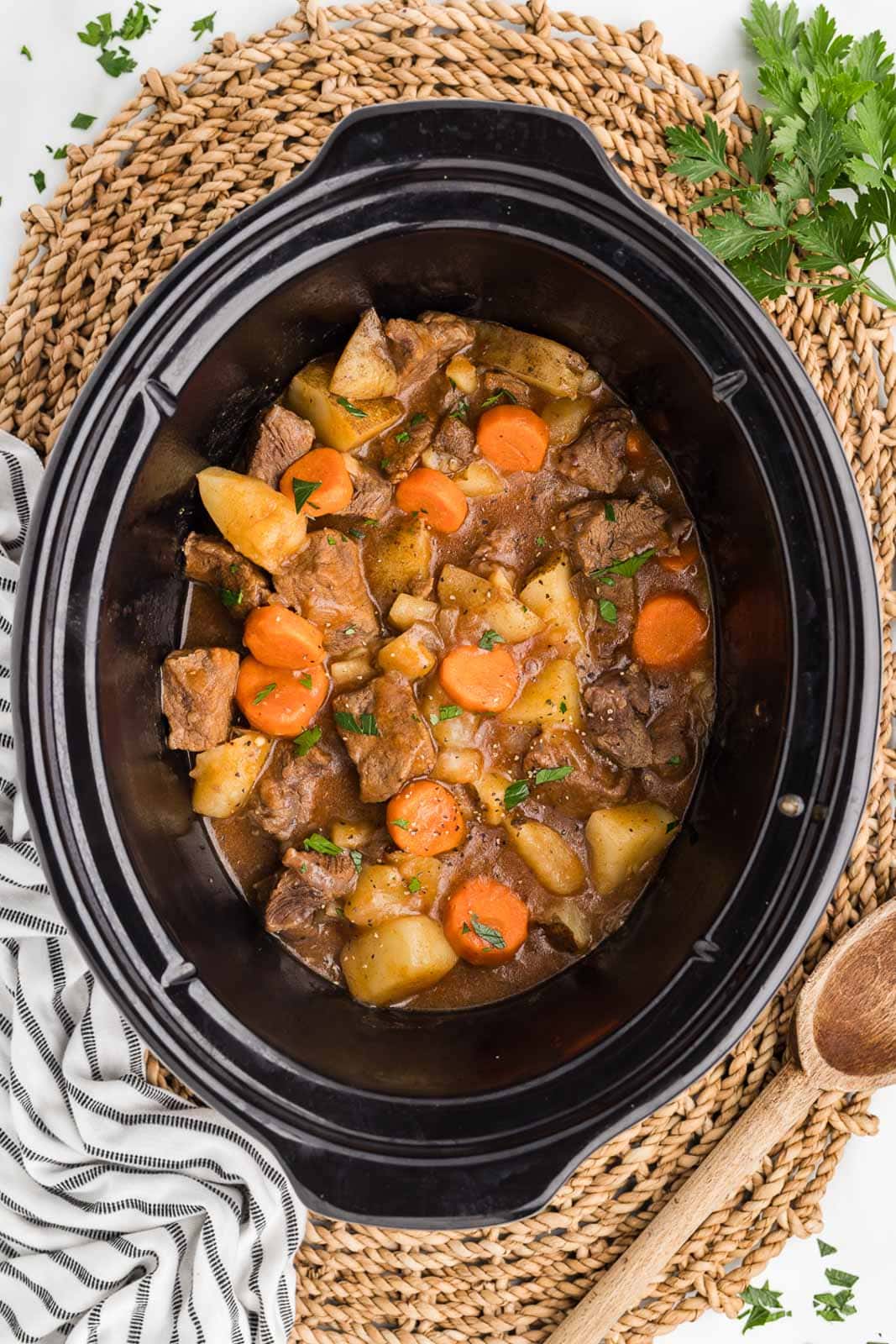 black slow cooker with cooked beef stew mixture lying on a woven food hot mat, with with and black striped dishcloth and wooden spoon on side