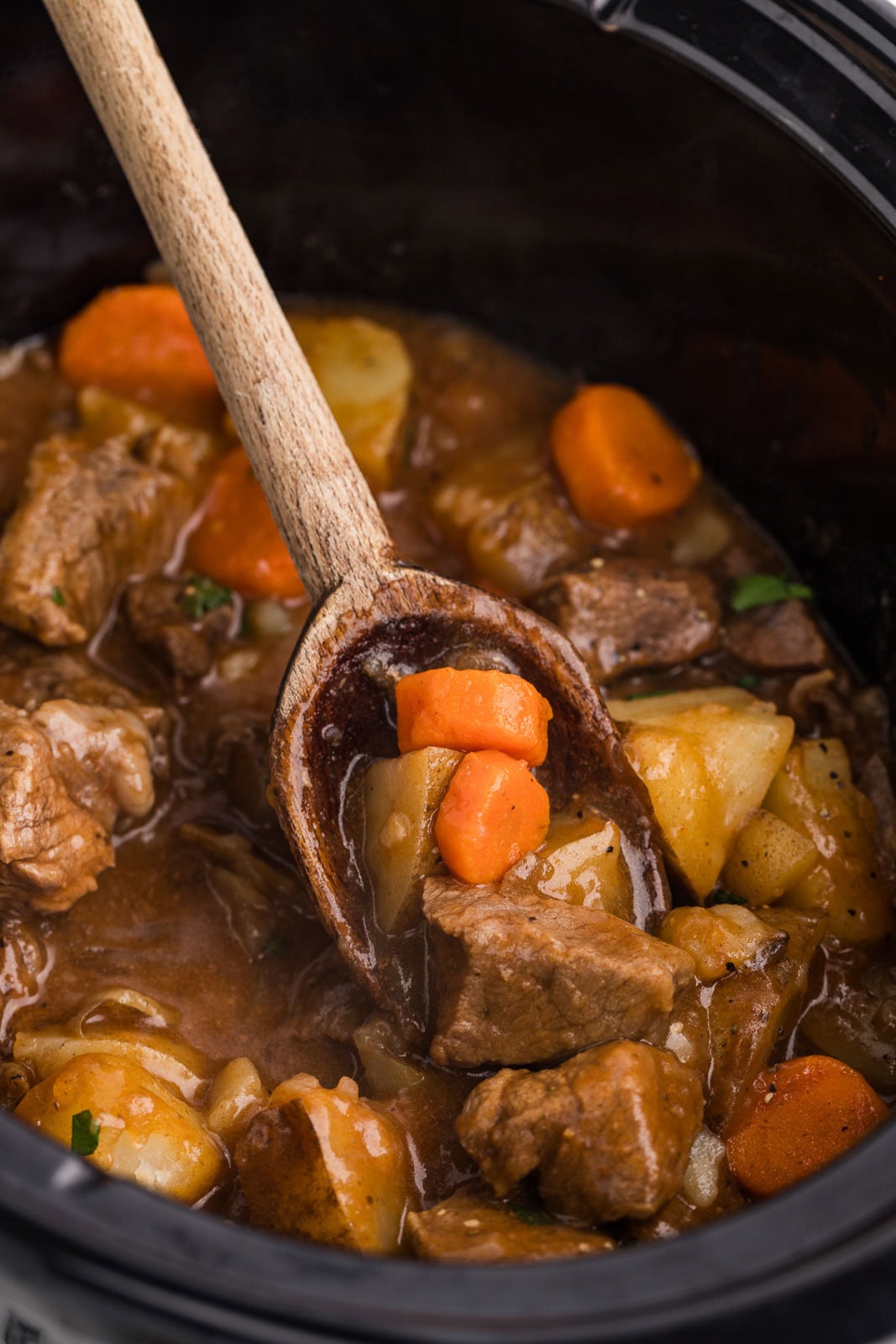closeup of black slow cooker containing beef stew with wooden spoon
