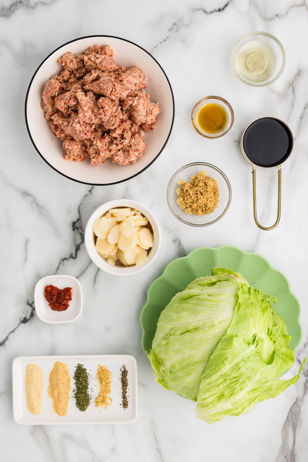 lettuce wraps ingredients to include white bowl of ground pork, green plate of head of lettuce, white plate with seasonings, containers of water chestnuts, soy sauce, fish sauce, rice vinegar, brown sugar and chili garlic sauce