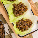 overhead view of wooden cutting board and white serving platter containing three lettuce leaves filled with pork mixture surrounded by bowl of sesame seeds and white bowl of pork mixture and chopsticks