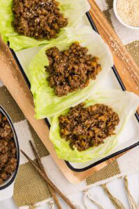 overhead view of wooden cutting board and white serving platter containing three lettuce leaves filled with pork mixture surrounded by bowl of sesame seeds and white bowl of pork mixture and chopsticks