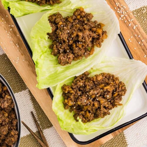 overhead view of wooden cutting board and white serving platter containing three lettuce leaves filled with pork mixture surrounded by bowl of sesame seeds and white bowl of pork mixture and chopsticks