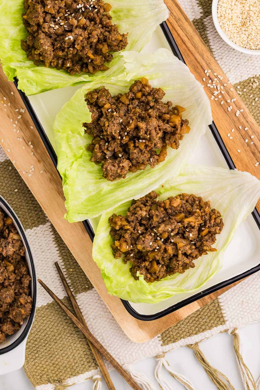 overhead view of wooden cutting board and white serving platter containing three lettuce leaves filled with pork mixture surrounded by bowl of sesame seeds and white bowl of pork mixture and chopsticks