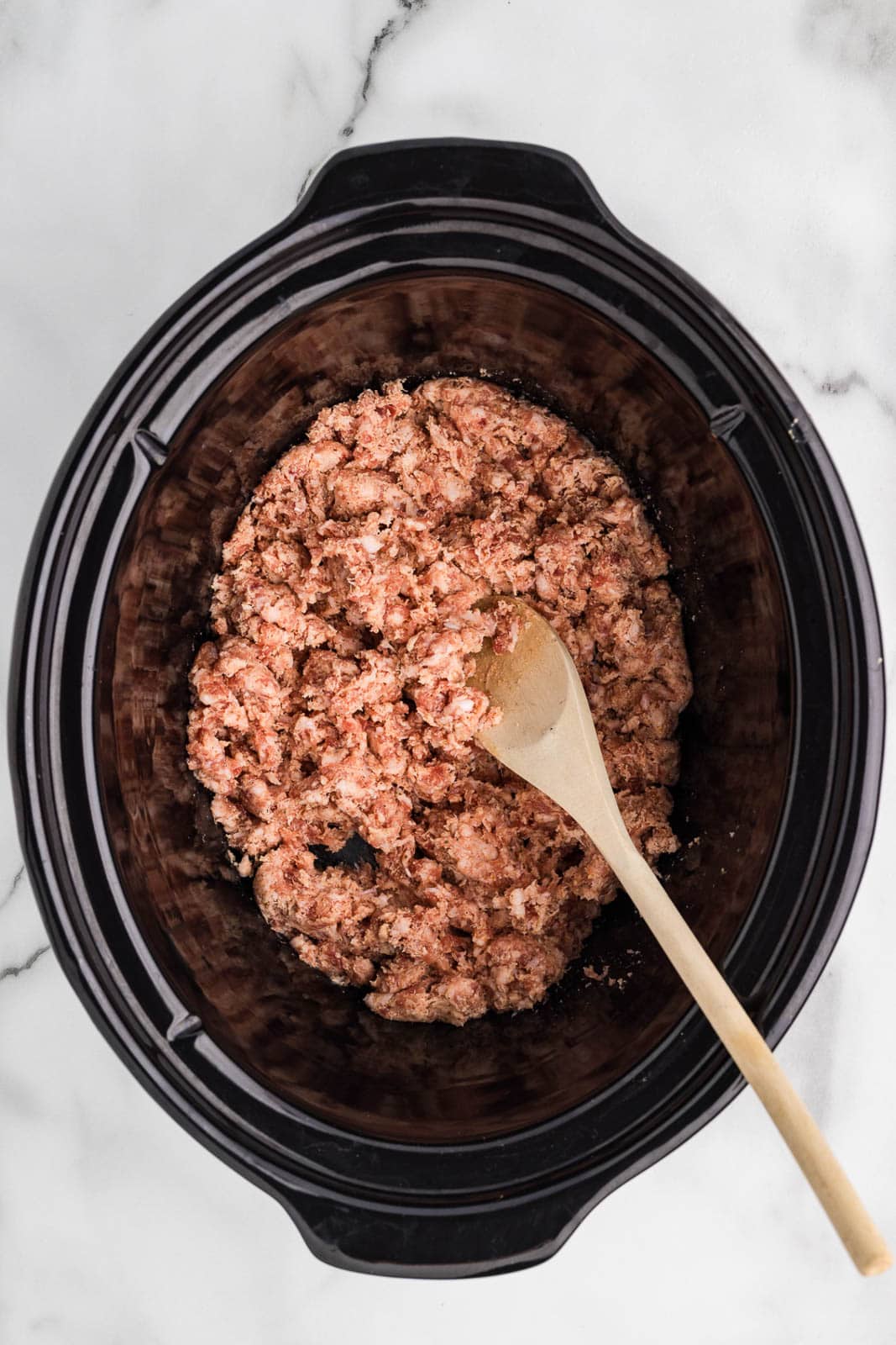 overhead view of ground pork in black slow cooker with wooden spoon