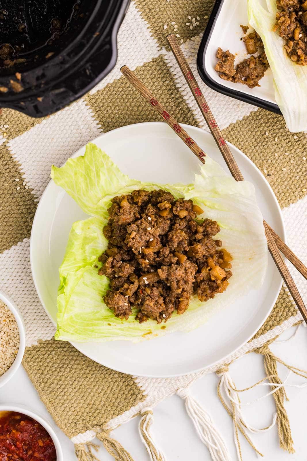 overhead view of white dinner plate with lettuce filled with pork mixture with two chopsticks, surrounded by black slow cooker, white serving platter, and two small white bowls of add ons