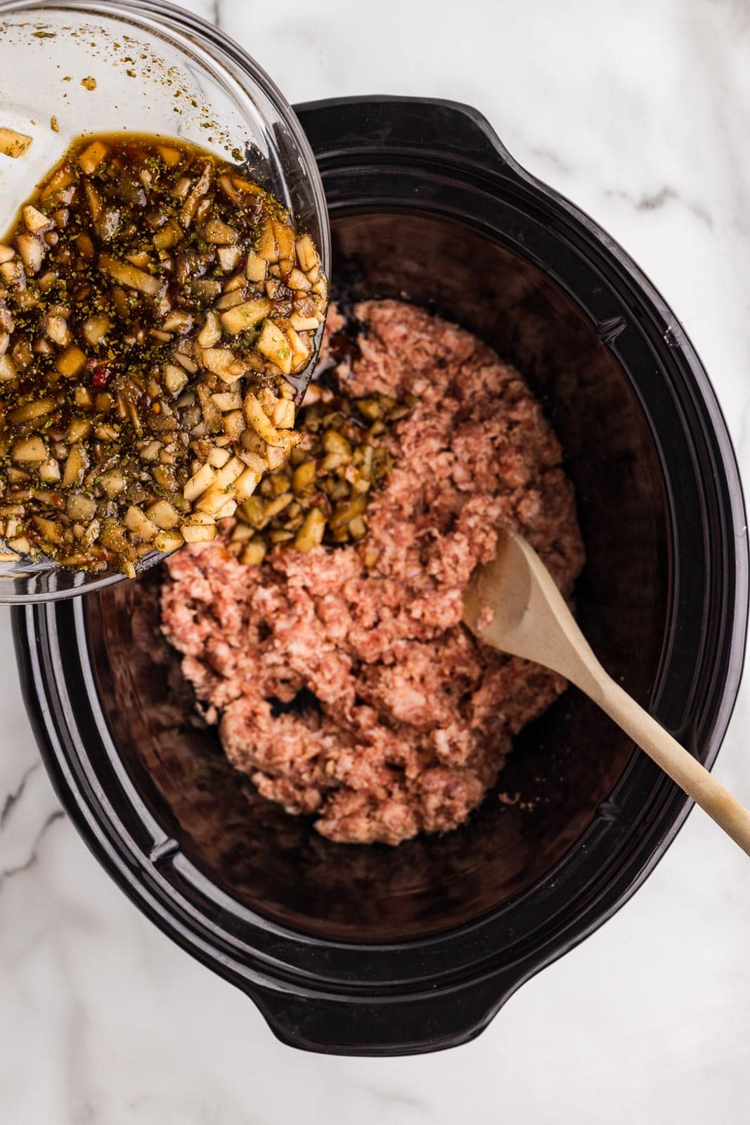 overhead view of black slow cooker with ground pork and wooden spoon with glass bowl of other ingredients being poured into slow cooker