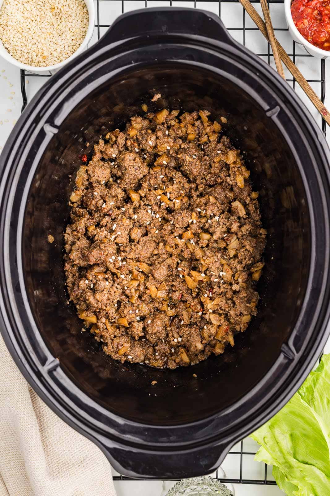 overhead view of slow cooked pork mixture in slow cooker surrounded by chopsticks, lettuce, tan dishcloth and two white bowls of other add ons