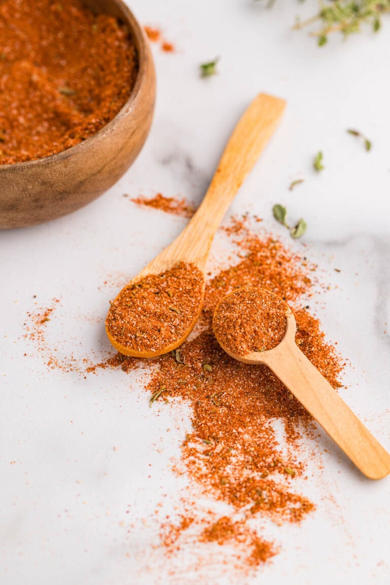 Small wooden measuring spoon and larger wooden spoon containing seasoning mixture with seasoning on surface and wooden bowl with seasoning in background