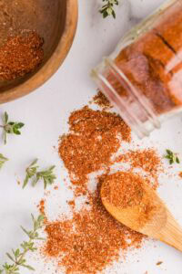 wooden bowl with small amount of seasonings, white glass container tipped over with seasonings on surface and wooden spoon