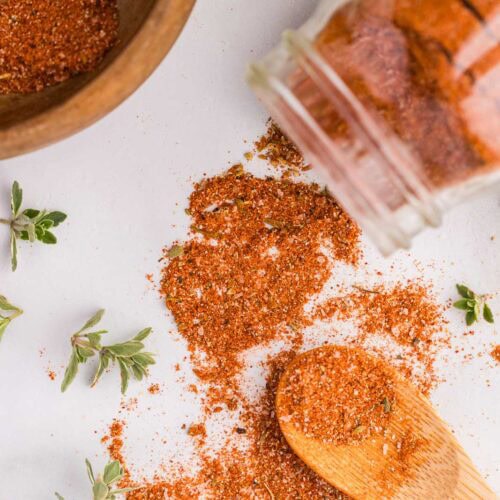 wooden bowl with small amount of seasonings, white glass container tipped over with seasonings on surface and wooden spoon