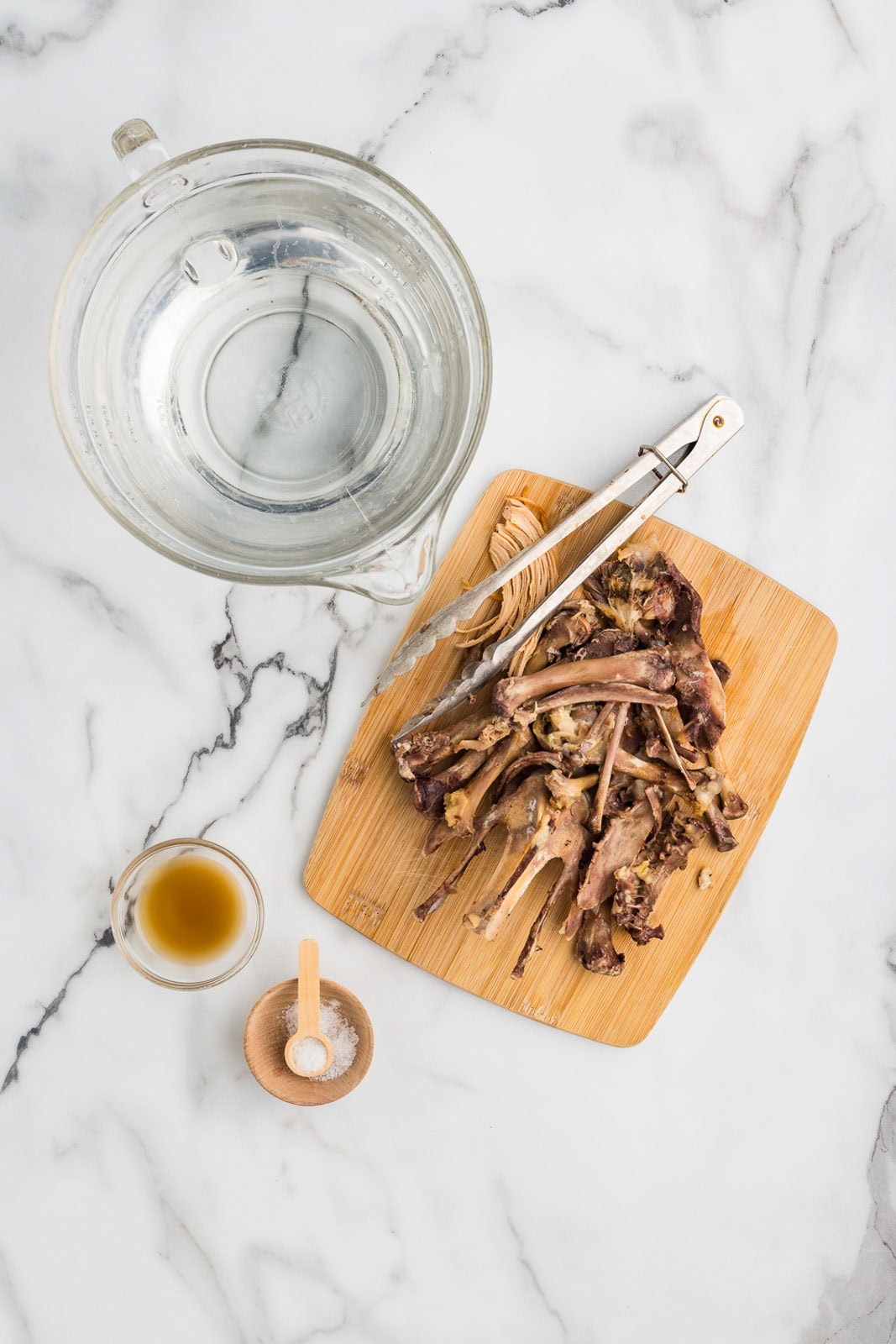 bone broth ingredients to include wooden cutting board of chicken bones with tongs, glass measuring cup of water, small glass bowl of apple cider vinegar and small wooden bowl of salt with measuring spoon