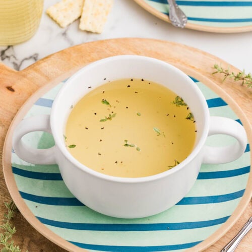 wooden cutting board containing blue and green striped plate with white cut of bone broth sprinkled with seasoning with spoon on side, with another plate, bowl and spoon, crackers and jar of bone broth in background