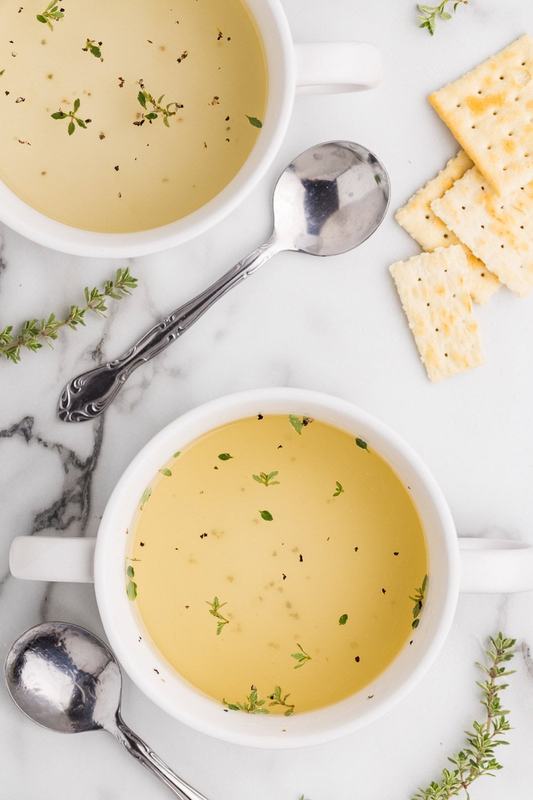 overhead view of two cups of bone broth sprinkled with seasoning surrounded by saltine crackers and two silver spoons