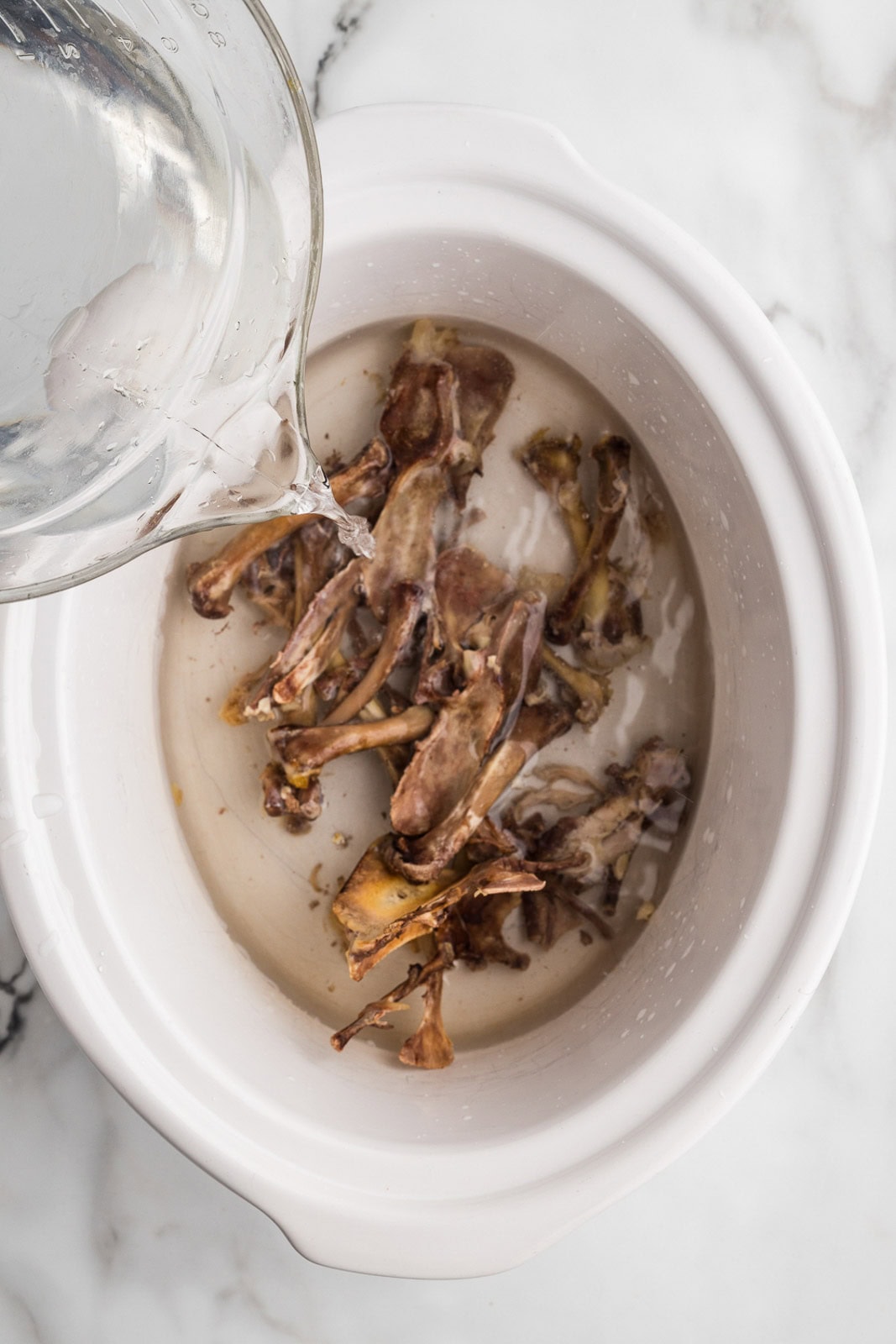 overhead view of white slow cooker containing chicken bones with glass measuring cup of water being poured into slow cooker