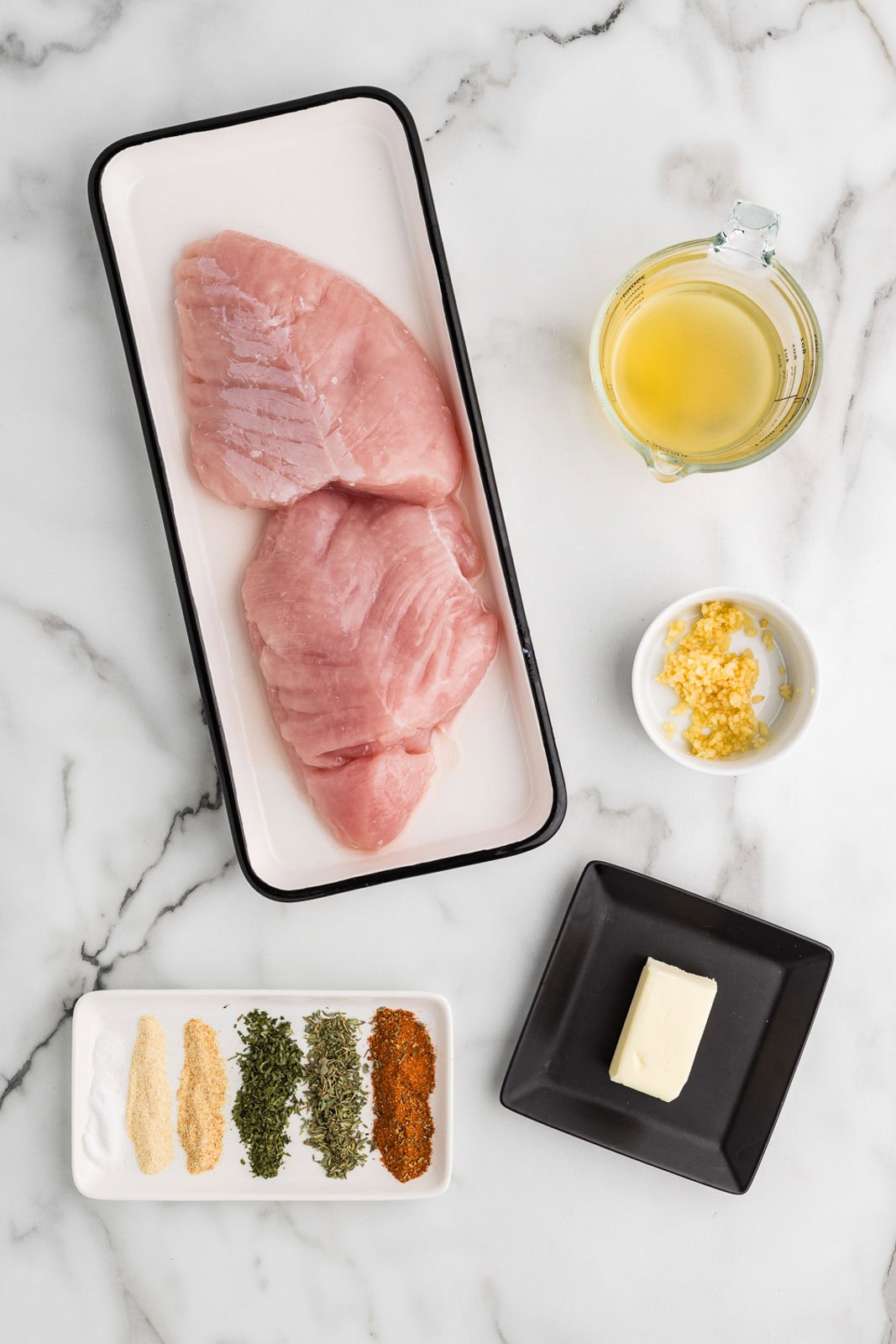 ingredients to include white platter with 2 pieces of turkey breast, glass measuring cup of chicken broth, bowl of minced garlic, black plate with butter, platter with seasonings