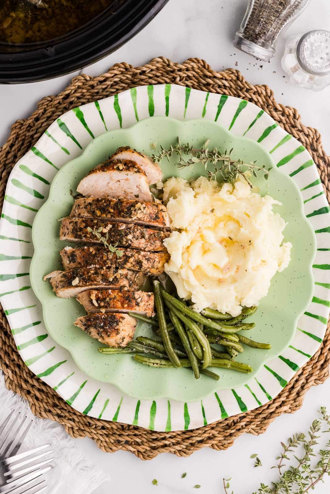 overhead view of light green plate on a mat containing sliced turkey breast, mashed potatoes and green bean, salt and pepper shakers, two forks and slow cooker in background