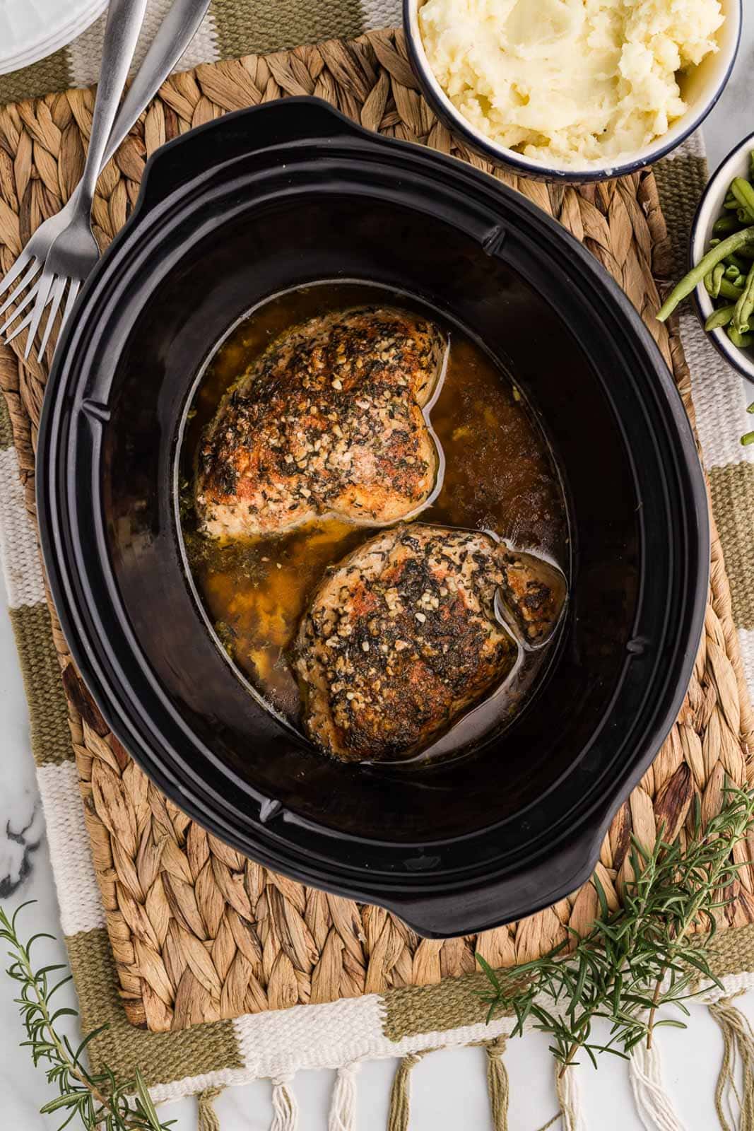 overhead view of black slow cooker containing slow cooked turkey breast on a mat surrounded by bowls of green beans, potatoes and 2 forks