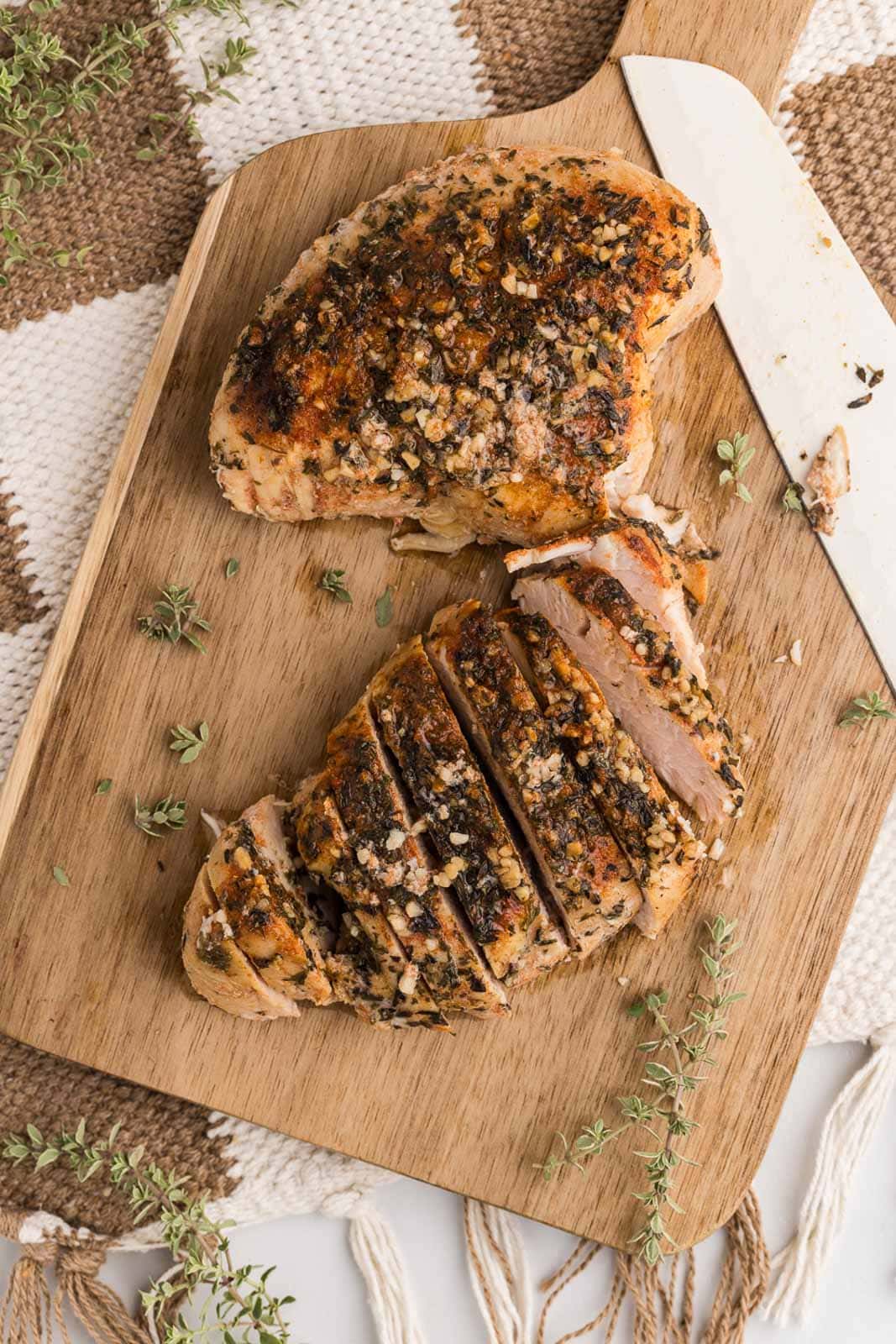 wooden cutting board on cloth mat with one sliced turkey breast and one not yet cut with a knife on the side