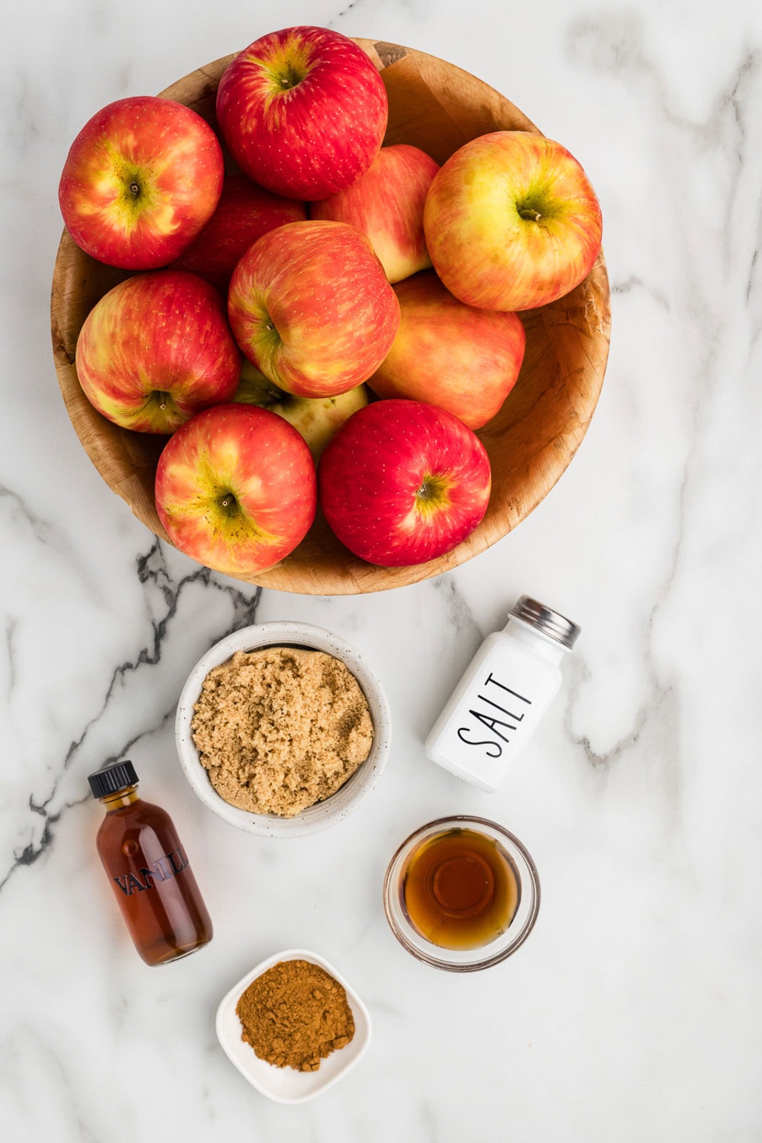 wooden bowl containing red apples surrounded by salt shaker white bowl of brown sugar, bowl of pumpkin pie spice, clear bowl of maple syrup and bottle of vanilla extract