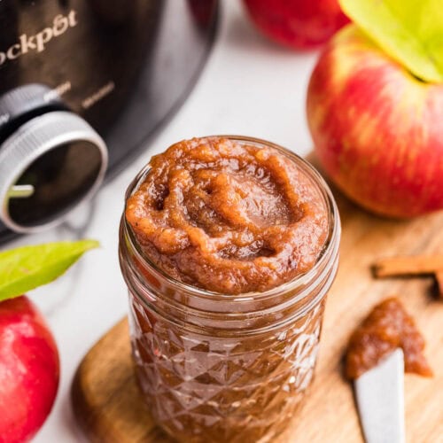 overhead view of wooden cutting board with glass jar of apple butter, silver knife, cinnamon sticks, surrounded by apples with black slow cooker in background