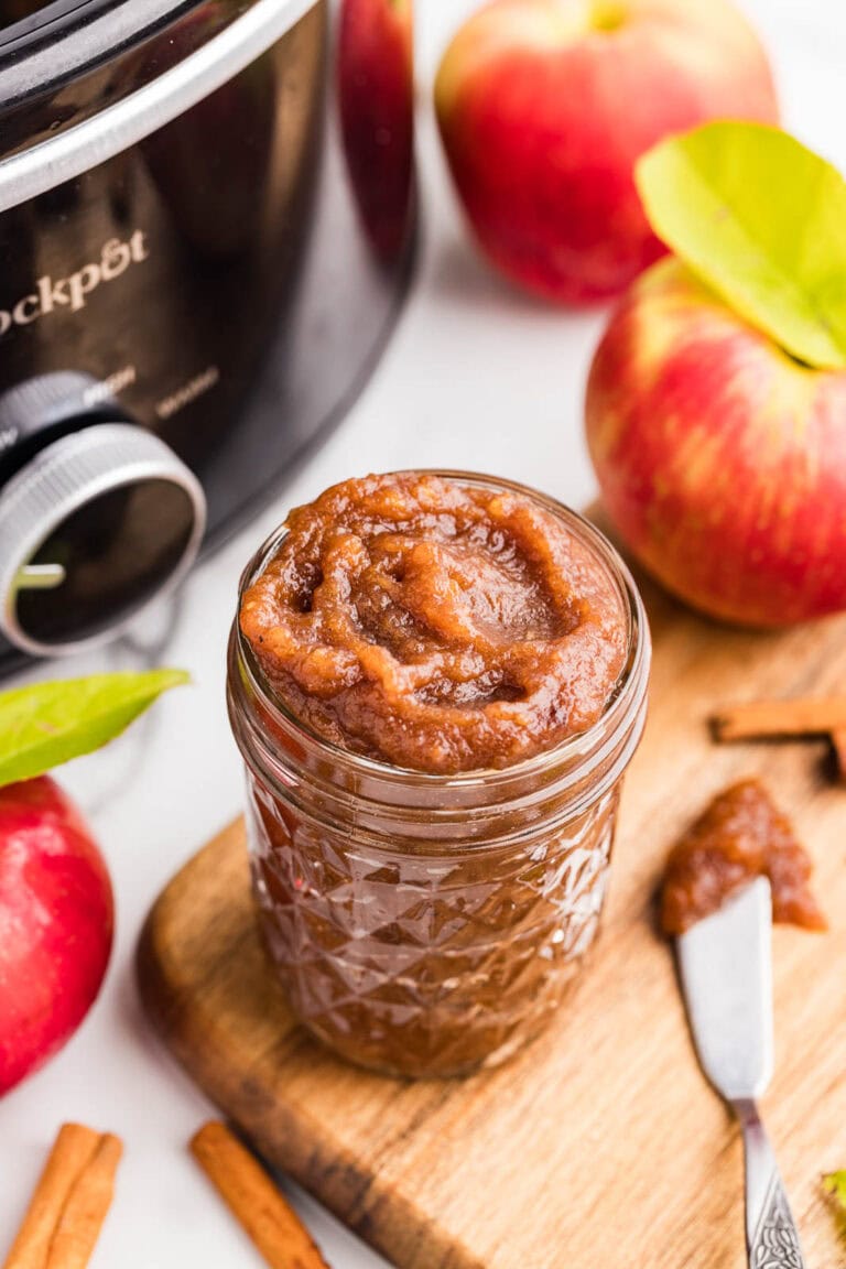 overhead view of wooden cutting board with glass jar of apple butter, silver knife, cinnamon sticks, surrounded by apples with black slow cooker in background