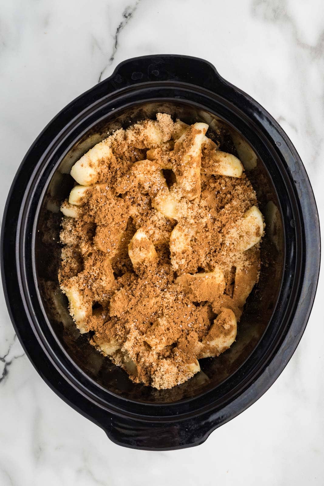 overhead view of black slow cooker with sliced apples covered by brown sugar