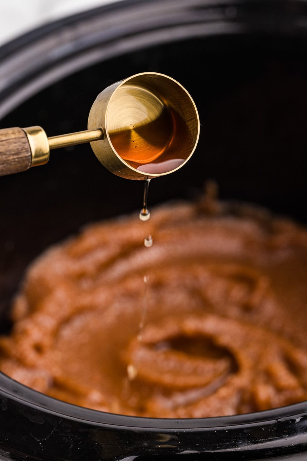 closeup of gold measuring spoon with wooden handle of syrup with black slow cooker with blended apples in background