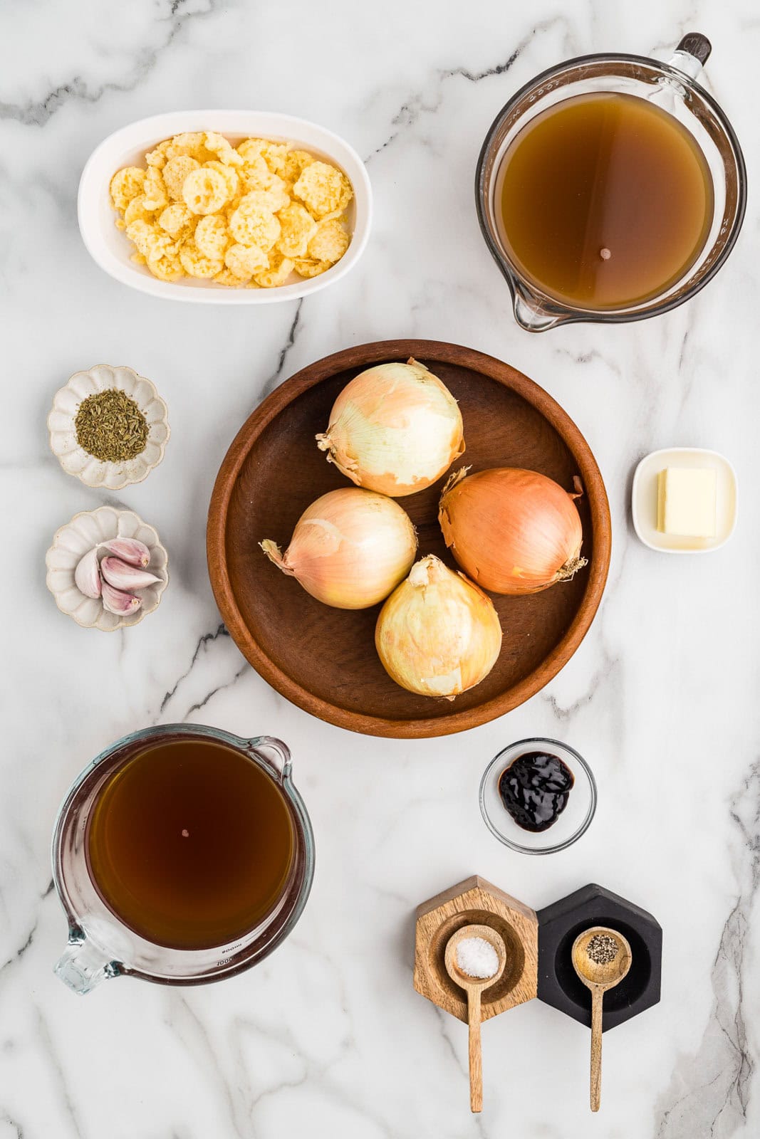 French onion soup ingredients to include wooden bowl with 4 onions, white bowl of Parmesan cheese crisps, glass measuring cups of beef broth and bone broth, small white containers of garlic cloves, butter, seasoning, glass bowl of balsamic glaze, and wooden bowls with spoon of salt and pepper