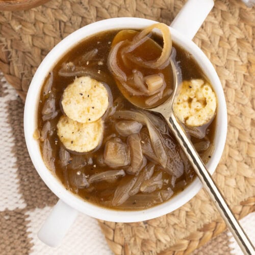 overhead view of white soup cup of onion soup and Parmesan crisps with silver spoon on a tan place mat with bowl of Parmesan crisps and salt and pepper shakers in the background
