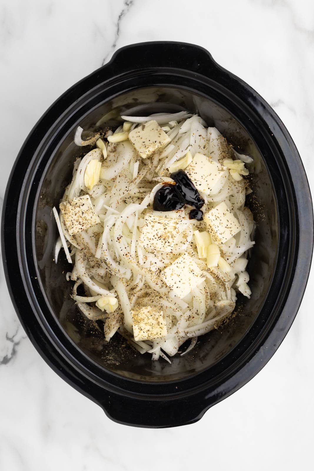 black slow cooker of sliced onions covered with seasonings with butter and balsamic glaze