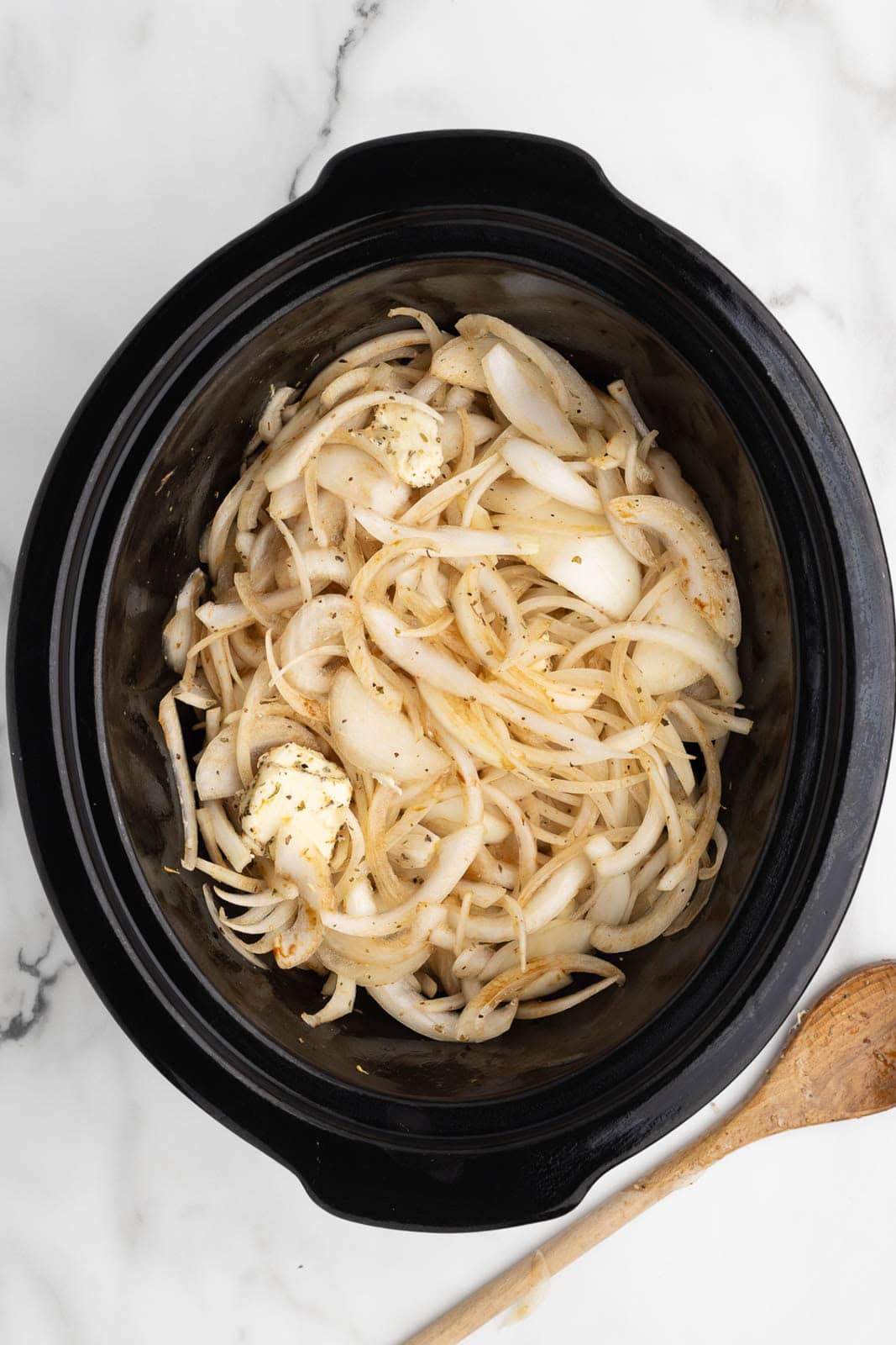 black slow cooker of sliced onions covered with seasonings with butter and balsamic glaze that have been stirred