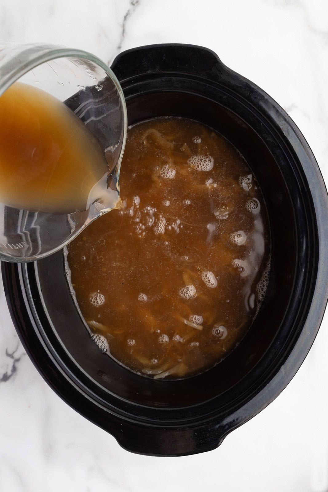 black slow cooker with onion mixture with broth and measuring cup of broth being poured into slow cooker