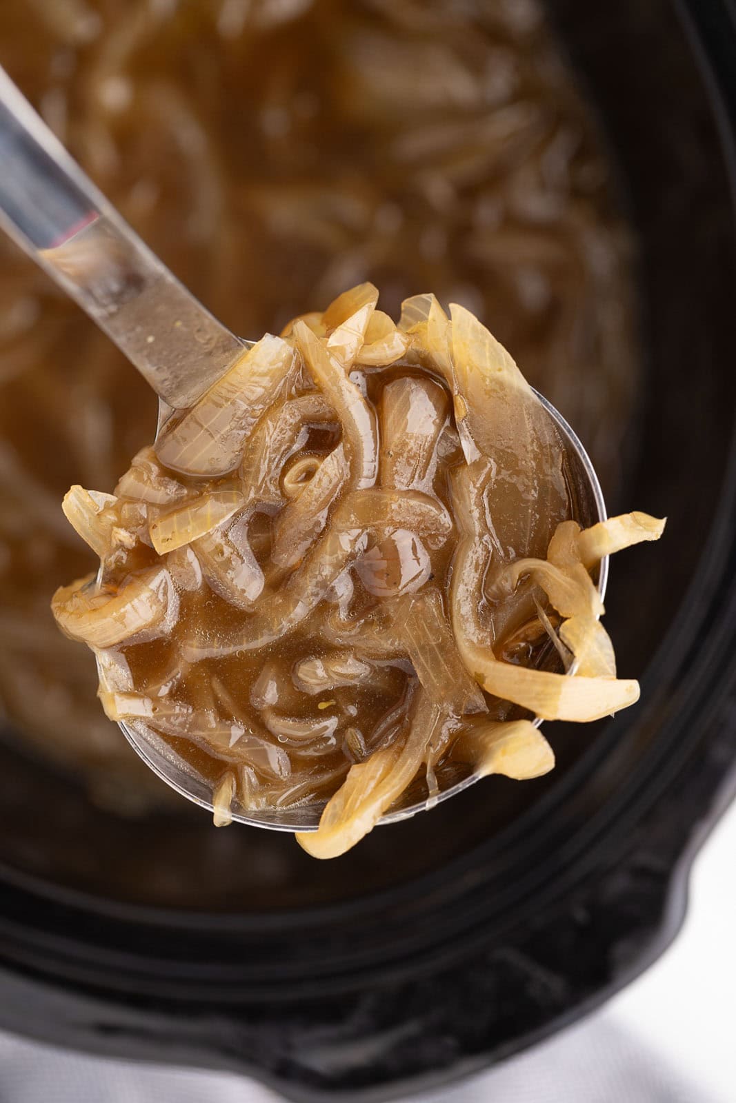 closeup view of large silver ladle of onion soup with black slow cooker of onion soup on background