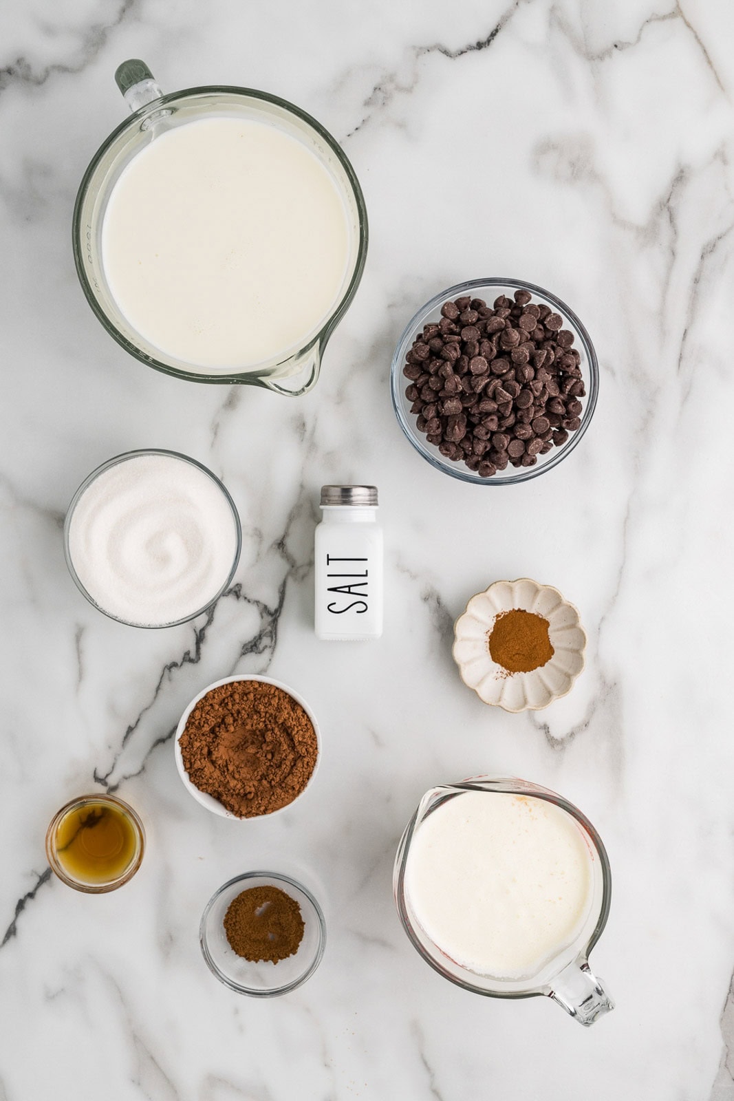 hot chocolate ingredients to include measuring cups of milk and heavy cream, bowls of cocoa powder, sugar, vanilla, espresso powder, ground cinnamon, chocolate chips and a salt shaker