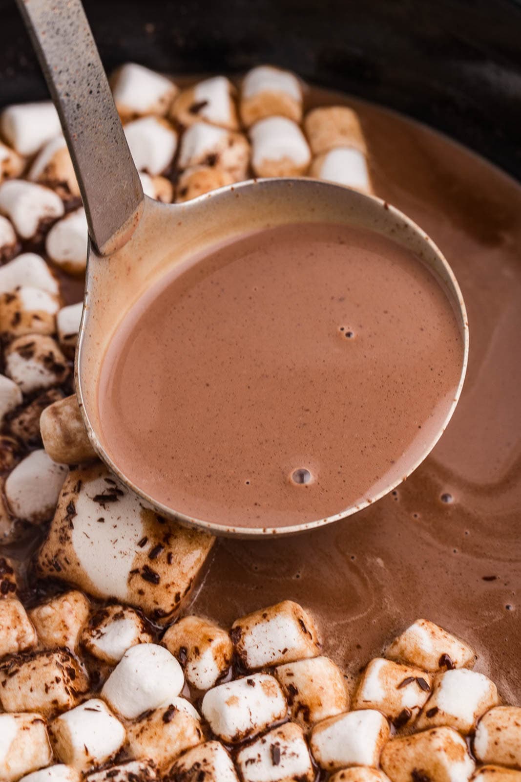 closeup view of ladle containing hot chocolate with slow cooker of mixture with marshmallow in background