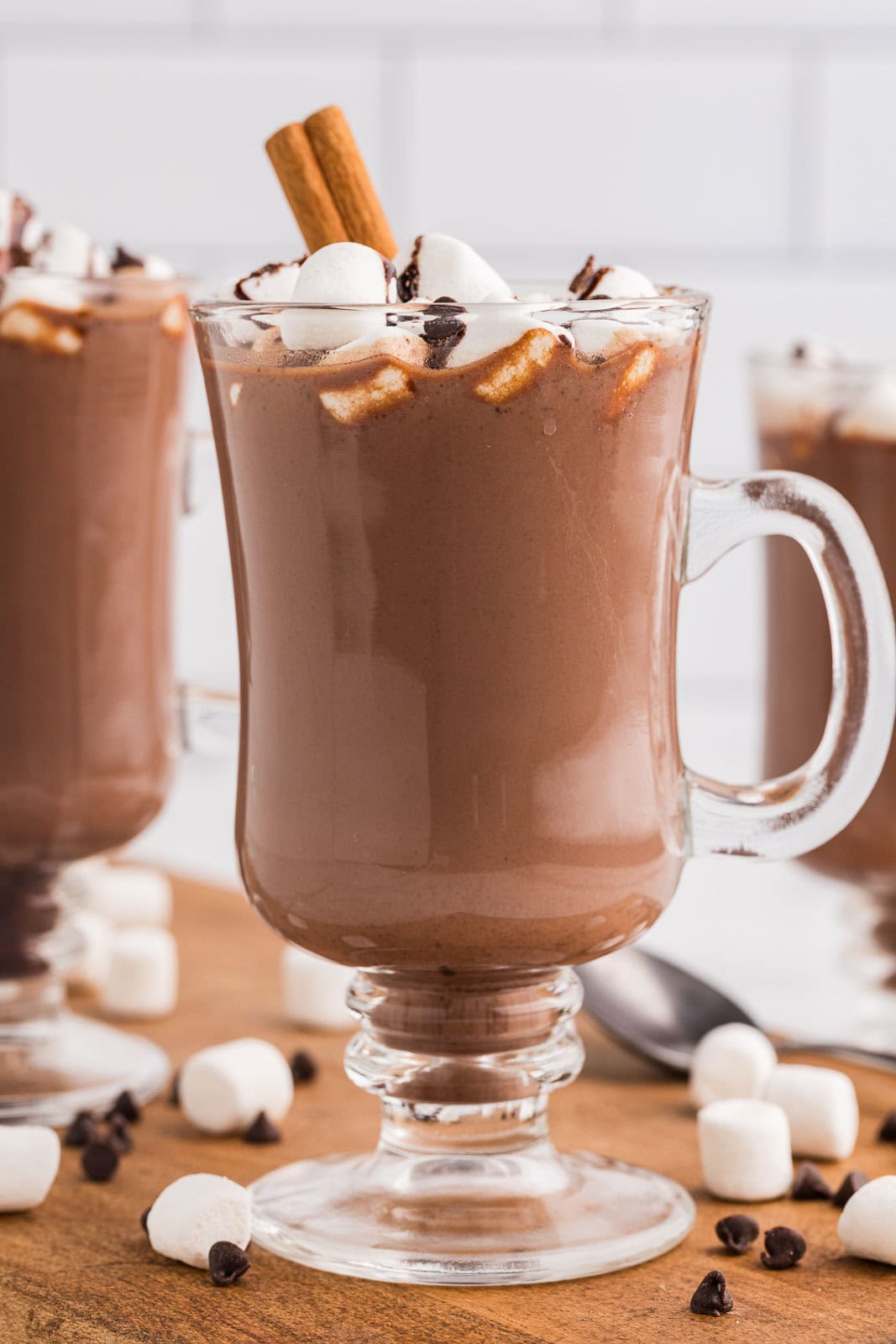 tall glass handled cup of hot chocolate with marshmallow and cinnamon sticks with 2 glasses in background and marshmallows and chocolate chips scattered on table