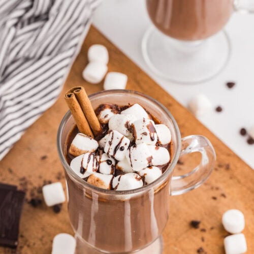 overhead view of glass containing hot chocolate with marshmallows and a cinnamon stick with another glass in background with scattered marshmallows on table