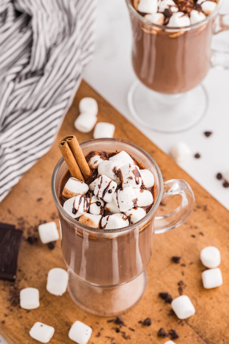 overhead view of glass containing hot chocolate with marshmallows and a cinnamon stick with another glass in background with scattered marshmallows on table