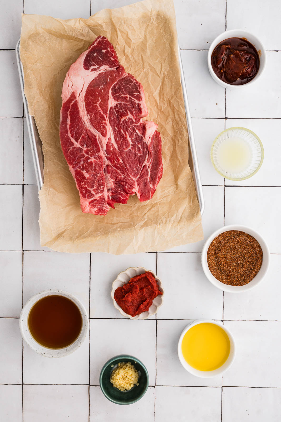 ingredients to include parchment paper lined sheet pan containing chuck roast, surrounded by small bowls of southwest spice blend, minced garlic, chipotle in adobo, orange juice, lime juice, beef broth and tomato paste