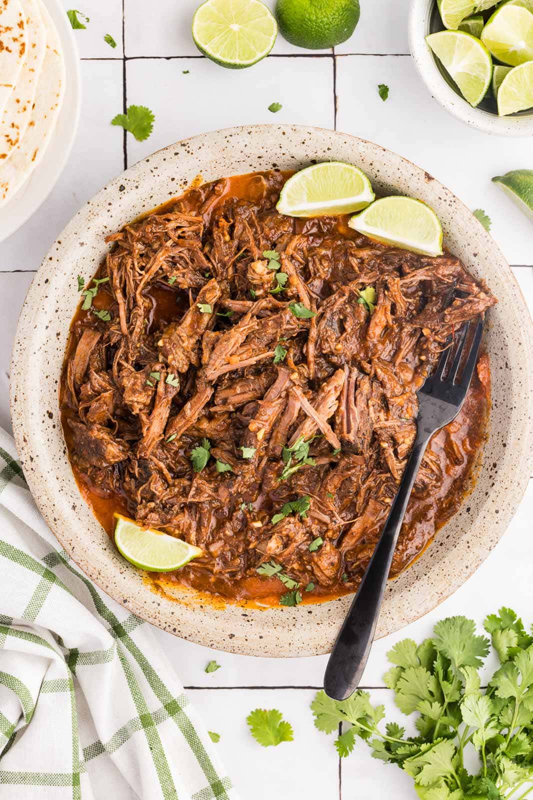 overhead view of speckled plate containing barbacoa with black forks and 3 limes, with limes, parsley and green and white striped dish towel surrounding plate