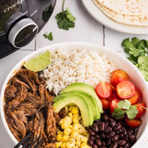 overhead view of white bowl containing Barbacoa beef, corn, black beans, cherry tomatoes. avocado and rice, surrounded by plate with tortillas, parsley, limes, green and white striped dish towel, black fork and black slow cooker